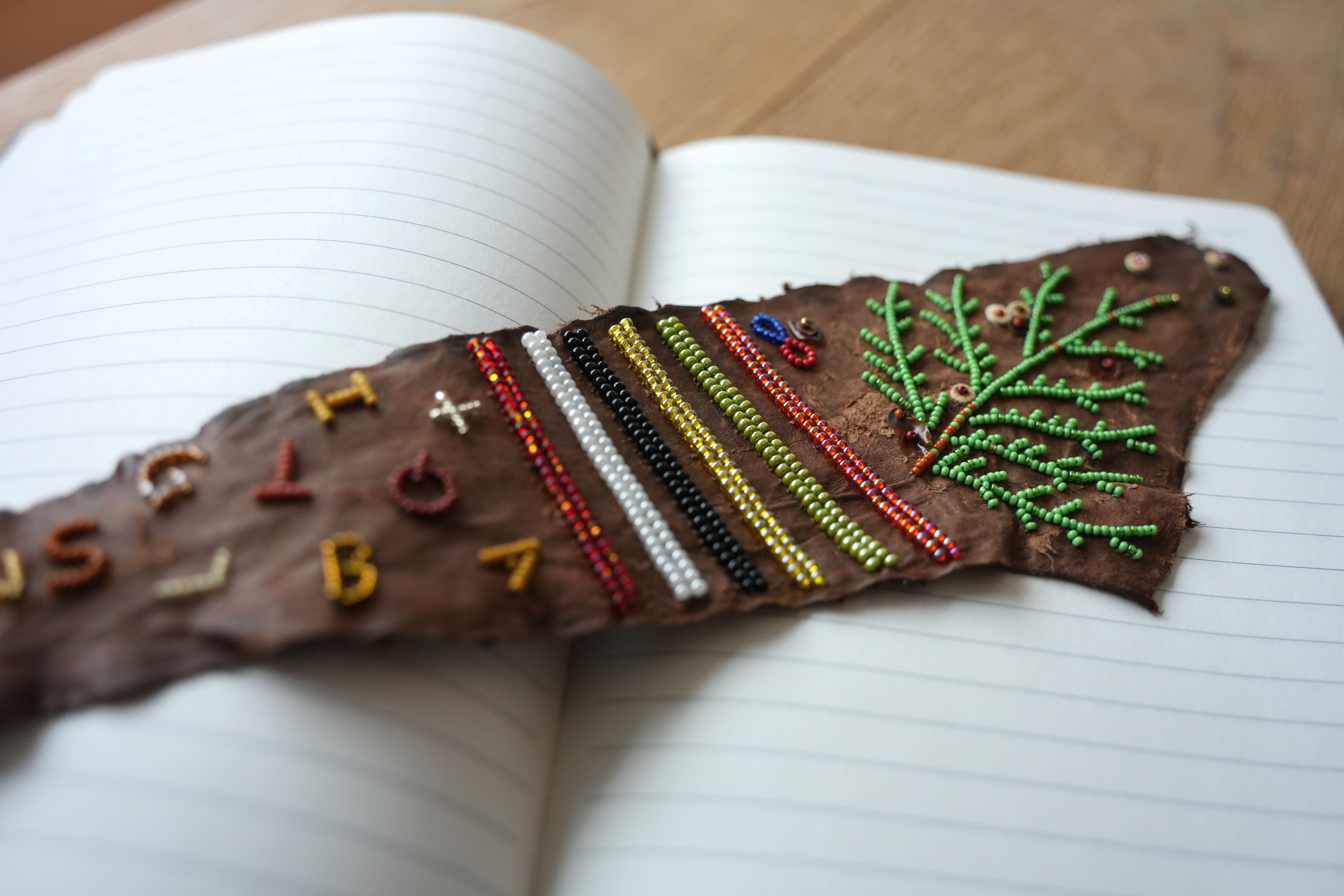 A photo of a piece of beadwork on fish skin leather, laying on top of an open book with lined pages. The beadwork includes letters, lines, and a pine tree branch.